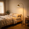Orsay Gold Arc Floor Lamp casting a soft warm glow over a cosy bedroom beside the bed