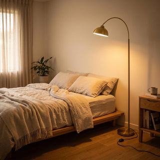 Orsay Gold Arc Floor Lamp casting a soft warm glow over a cosy bedroom beside the bed