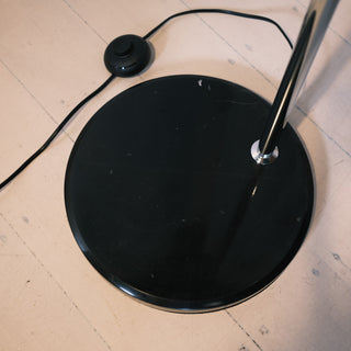 A high-angle view of the chrome arc floor lamp's heavy, round black base resting on light wooden floorboards. A convenient black foot switch is visible on the power cord, demonstrating the lamp's practical design and stability.