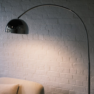 The chrome arc floor lamp with its domed shade illuminated, casting a bright, functional light over a cream lounge chair. It stands beside a round wooden side table stacked with design books and a plant, set against a backdrop of white brick and sheer curtains.