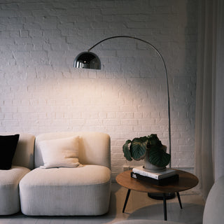 A cozy lounge setting featuring the chrome arc floor lamp casting a warm glow over a cream sofa. A plant and a stack of books rest on a nearby side table, completing the relaxed yet stylish atmosphere of the room.