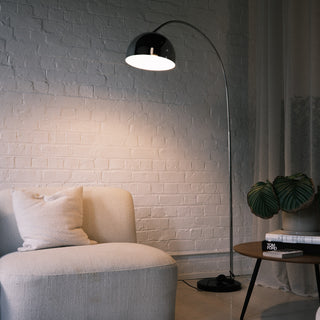 A cozy reading corner illuminated by a chrome arc floor lamp featuring a domed shade. It hovers over a cream sofa with a cushion, next to a side table stacked with design books and a plant, creating a warm ambient glow against the textured wall.