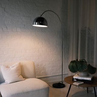 A modern chrome arc floor lamp arching elegantly over a cream modular sofa, providing overhead lighting. The lamp stands next to a wooden round table with a large potted plant, set against a white brick wall in a minimalist living space.