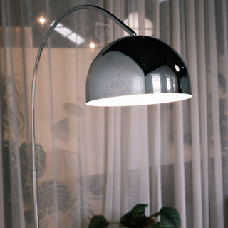 A close-up focus on the highly polished chrome domed shade of the arc floor lamp. The mirror-like metal surface reflects the surroundings, while the downward light casts a warm, ambient glow across the textured white brick wall, highlighting the sleek contrast between the modern fixture and the rustic background.