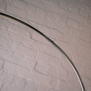 A close-up detail shot focusing on the smooth, curved silver stem of the chrome arc floor lamp. The image highlights the high-quality polished metal finish and the seamless connection points against a textured white brick wall.