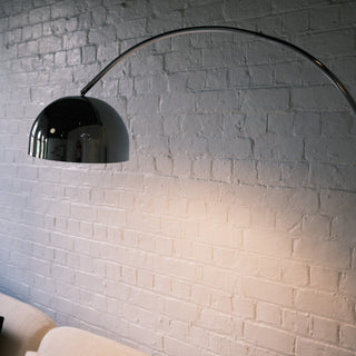 A close-up detail shot of the chrome arc floor lamp, highlighting its polished, reflective domed shade against a painted white brick wall. The curve of the silver stem is visible, emphasizing the sleek, modern design and the quality of the finish.