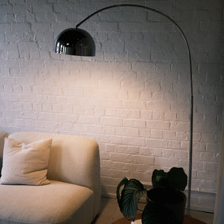 The chrome arc floor lamp displayed in an entryway setting beside a dark wood console table. Rustic wooden log stools and a large textured floor vase complement the modern silver finish of the lamp against the white brick background.