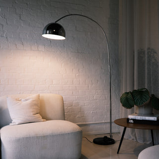 A modern chrome arc floor lamp featuring a polished domed shade, arching elegantly over a cream armless chair. The lamp stands beside a round wooden side table with a potted plant, set against a textured white brick wall that enhances the minimalist aesthetic.