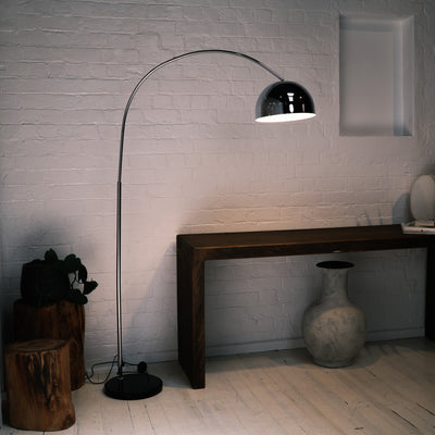 The chrome arc floor lamp positioned beside a dark wood console table, highlighting a blend of modern and rustic styles. Wooden stump stools and a large textured vase sit on the floor, while the lamp’s polished finish stands out against the white brick wall.