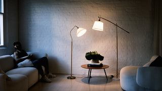 A warm living room scene featuring two designer floor lamps, one pleated shade lamp and one arc lamp, styled between two modern sofas with a small coffee table. Soft ambient lighting highlights the brick wall and cosy lounge setting.