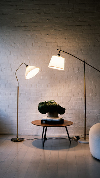 A mobile view of two designer floor lamps beside a wooden coffee table with a plant, set against a textured white brick wall and soft ambient lighting.
