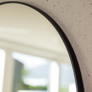 Close-up of the Grandeur X arched mirror’s curved top edge showing the smooth black aluminium frame and precise contour against a textured wall surface. The reflection captures soft natural light from a bright modern interior, emphasising the mirror’s elegant arched shape and fine craftsmanship.