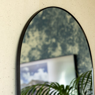Close-up of the Grandeur X arched mirror frame showing its smooth black aluminium edge and fine craftsmanship. The reflection reveals part of a wall artwork and indoor plant, highlighting the mirror’s clean curve and modern finish.
