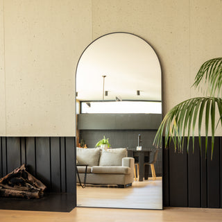 A 200 × 100 cm Grandeur X arched full-length mirror with a slim black frame standing beside a modern fireplace and indoor palm. The mirror reflects a cosy living space with a beige sofa, wooden flooring and black wall panels, highlighting its large, elegant shape and ultra-clear reflection.
