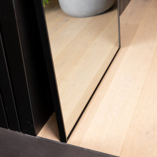 Close-up of the Grandeur X arched mirror base resting on a light timber floor beside a black wall panel. The image highlights the slim black aluminium frame and precise edge alignment, showing how seamlessly the mirror sits flush against the wall and floor.
