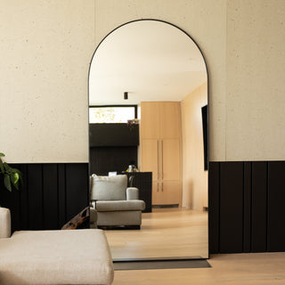 A large 200 × 100 cm arched full-length mirror with a slim black frame reflecting a bright, modern living area. The mirror stands against a textured wall with black panelling and light oak flooring, showing a soft grey armchair and warm wooden cabinetry in the background.