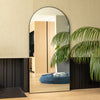 A tall arched full-length mirror with a slim black frame placed beside a light-toned wall and indoor palm plant. The mirror reflects a modern living space with warm oak flooring and soft natural light, showcasing the mirror’s clear reflection and elegant shape.