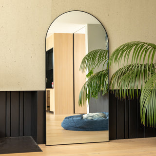 A tall arched full-length mirror with a slim black frame placed beside a light-toned wall and indoor palm plant. The mirror reflects a modern living space with warm oak flooring and soft natural light, showcasing the mirror’s clear reflection and elegant shape.