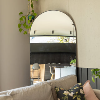 The Grandeur X arched full-length mirror placed neatly in the corner of a modern living room. The mirror reflects a bright open-plan kitchen with wooden tones, black tiles, and indoor plants, adding depth and calm to the space. A beige sofa with floral cushions sits in front, creating a warm and relaxed atmosphere.