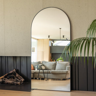 A 200 × 100 cm Grandeur X extra large arched mirror with a slim black frame placed beside a modern fireplace and indoor palm. The mirror reflects a bright living space with a beige sofa, black coffee table, and warm wooden tones, highlighting the mirror’s tall curved design and crystal-clear reflection.