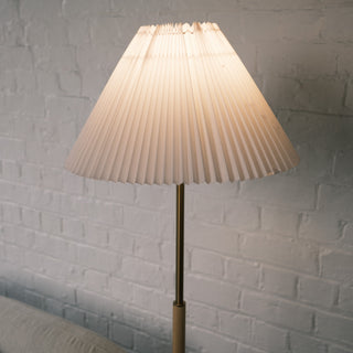 Close up of the Luma Pleated Floor Lamp’s soft white pleated shade, glowing warmly against a textured white brick wall.