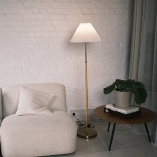 Luma Pleated Floor Lamp standing beside a cream sofa and wooden side table with a plant, softly lighting a white brick wall in a calm, modern living room.