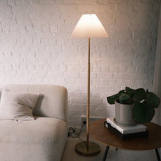 Luma Pleated Floor Lamp placed beside a cream fabric sofa and wooden side table with plants, softly illuminating a white brick wall in a modern living room.