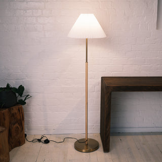 A full view of the Luma Pleated Shade Floor Lamp placed beside a wooden console table and indoor plant. The lamp features a white pleated tapered fabric shade, a warm brass pole, and a round weighted brass base. The soft light spreads gently across the white brick wall, creating a calm and inviting interior atmosphere.