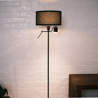 Front view of the Lumen Duo Floor Lamp showing the black fabric drum shade and the adjustable reading light below, standing against a white brick wall with warm ambient lighting.