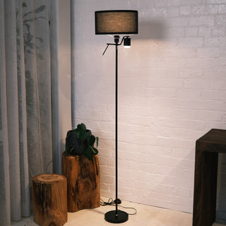 Black Lumen Duo Floor Lamp featuring a fabric drum shade and adjustable reading light, shown in a modern living room with white brick walls, neutral sofa, wooden side decor, and soft ambient lighting.