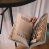 A person holding an open book under the focused reading light of the Lumen Duo Floor Lamp, with the lamp’s arm and shade subtly visible in the background.