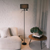 The Lumen Duo Floor Lamp standing next to a cream sofa and a wooden side table with a leafy plant, adding warm accent lighting to a calm, modern living room.