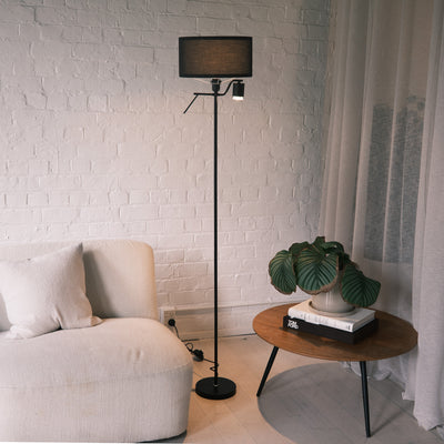 The Lumen Duo Floor Lamp standing next to a cream sofa and a wooden side table with a leafy plant, adding warm accent lighting to a calm, modern living room.