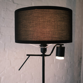 Close up of the Lumen Duo Floor Lamp showing its black fabric drum shade and the focused reading light beneath, set against a softly textured white brick wall.