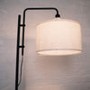 Side angle view of the Madox Loft standing lamp showing its curved black frame and warm fabric drum shade, styled against a textured white brick wall. Ideal for floor lamps NZ, standing lamps, modern floor lamps and dimmable lamps. | C&F Creation NZ