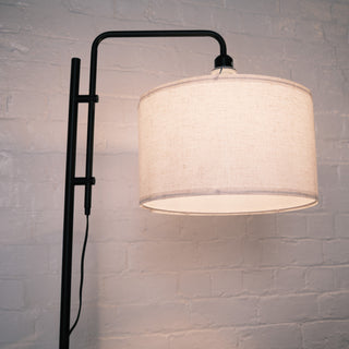 Side angle view of the Madox Loft standing lamp showing its curved black frame and warm fabric drum shade, styled against a textured white brick wall. Ideal for floor lamps NZ, standing lamps, modern floor lamps and dimmable lamps. | C&F Creation NZ