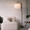 Modern floor lamp with a fabric drum shade and black metal frame placed beside a soft cream lounge chair in a minimalist NZ living room. A warm ambient lighting scene showcasing floor lamps, standing lamps, modern floor lamps and dimmable lamps. | C&F Creation NZ