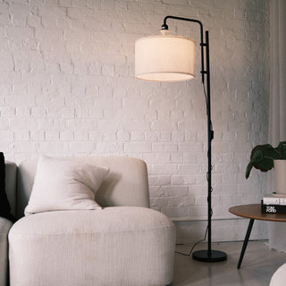 Modern floor lamp with a fabric drum shade and black metal frame placed beside a soft cream lounge chair in a minimalist NZ living room. A warm ambient lighting scene showcasing floor lamps, standing lamps, modern floor lamps and dimmable lamps. | C&F Creation NZ