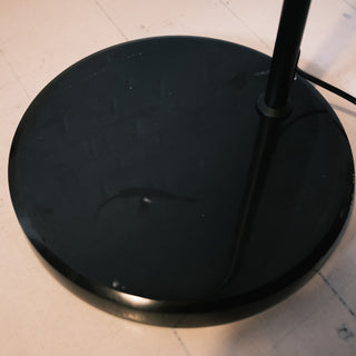 A close-up view shows the solid, round black metal base of the Nero floor lamp. It sits firmly on a light wooden floor. The smooth surface provides stable support for the structure.