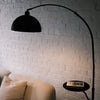 This Nero floor lamp has a smooth metal dome shade and a built-in tray. It sits beside a textured fabric armchair. Warm light reflects off the black steel frame.