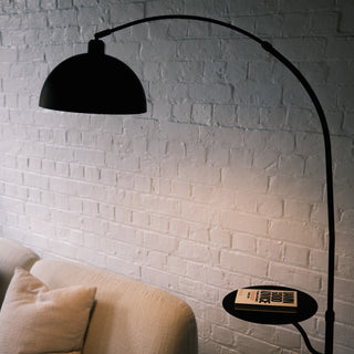 This Nero floor lamp has a smooth metal dome shade and a built-in tray. It sits beside a textured fabric armchair. Warm light reflects off the black steel frame.