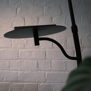A close-up highlights the circular metal tray and curved arm. A book rests on the matte black shelf. The solid steel construction stands out against a white brick wall.