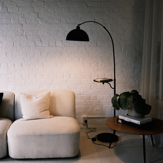 The Nero floor lamp features a black metal arched stem and a sturdy tray. It stands next to a soft fabric sofa. The matte finish contrasts with the wooden side table.