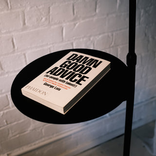 A close-up view shows the black dome shade and arched stem of the Nero floor lamp. A small book rests on its integrated tray. The background is a white brick wall.