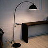 The Nero floor lamp features a black arched stem and a round tray. It stands beside a dark wooden console. A white brick wall creates a clean, modern room.