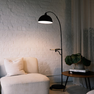 The Nero floor lamp features a black arched stem and a built-in tray. It sits beside a cream sofa. Soft curtains and a white brick wall create a warm, lit space.