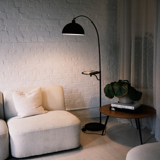The Nero floor lamp features a black arched stem and a built-in tray. It sits beside a cream sofa. Soft curtains and a white brick wall create a warm, lit space.