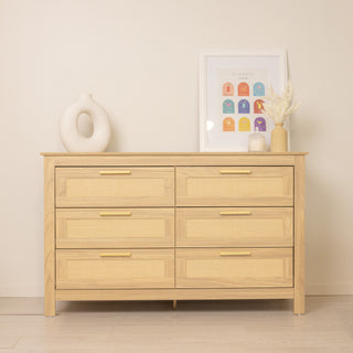 Raya 6 Drawer dresser in oak finish with rattan front panels and brass gold handles, styled with a circular ceramic vase, framed artwork, and a small dried floral arrangement in a light modern interior.