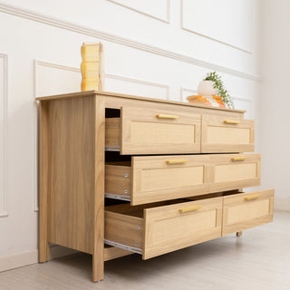 Raya 6-Drawer Lowboy dresser, 123 × 40 × 75 cm, in warm oak finish with rattan-style drawer fronts and gold handles, styled in a bright modern room with drawers open to show storage depth.