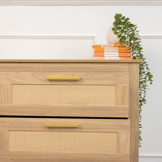 Close-up of the top right section of the Raya 6 Drawer dresser in oak finish with brass handles and rattan drawer fronts, styled with stacked orange books and a trailing green plant.